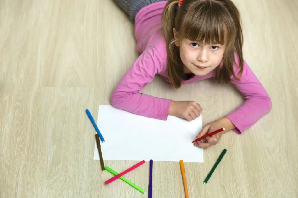 A little girl laying on the floor with colored pencils.